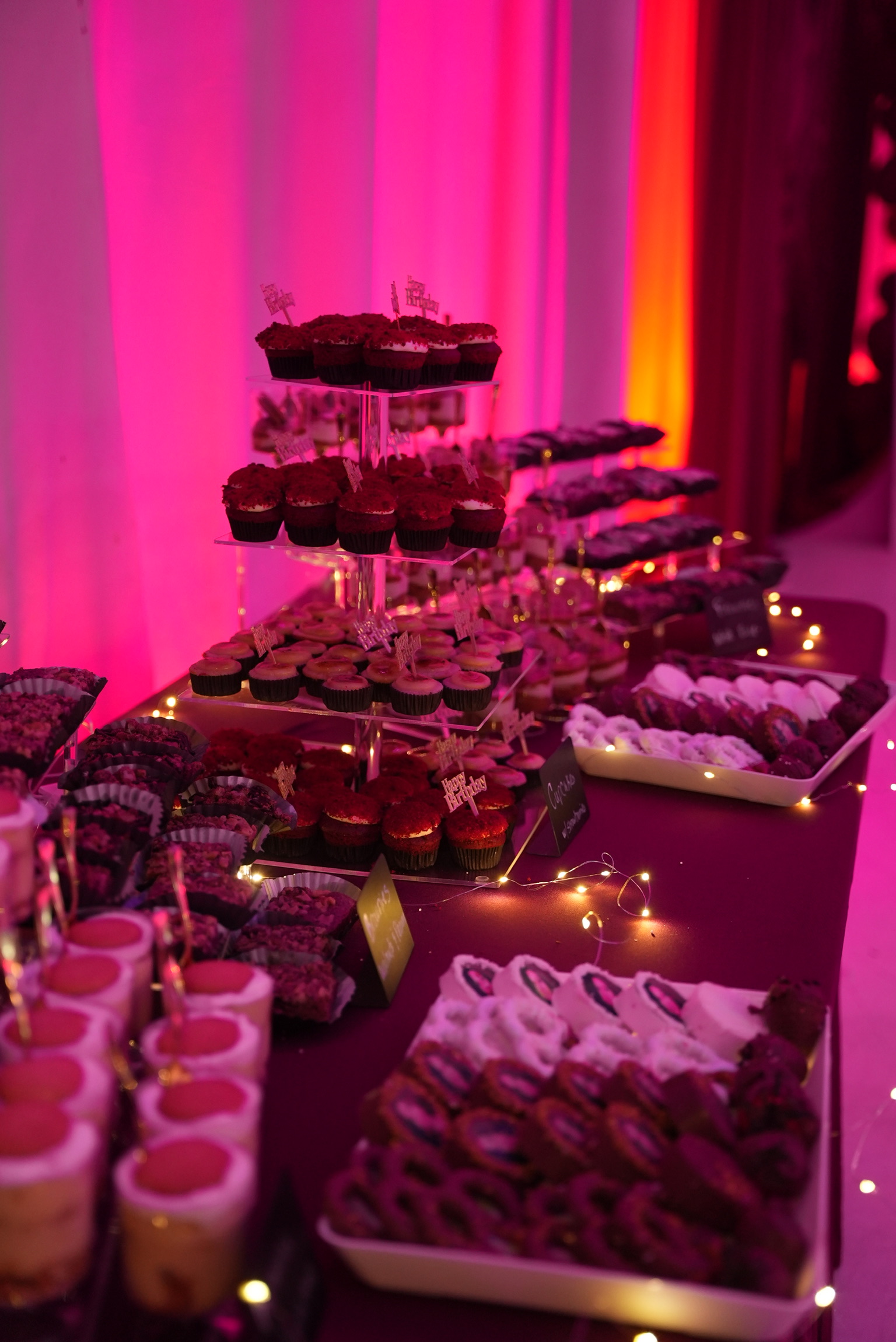 Birthday dessert table with cupcakes and an assortment of pastries under pink lighting