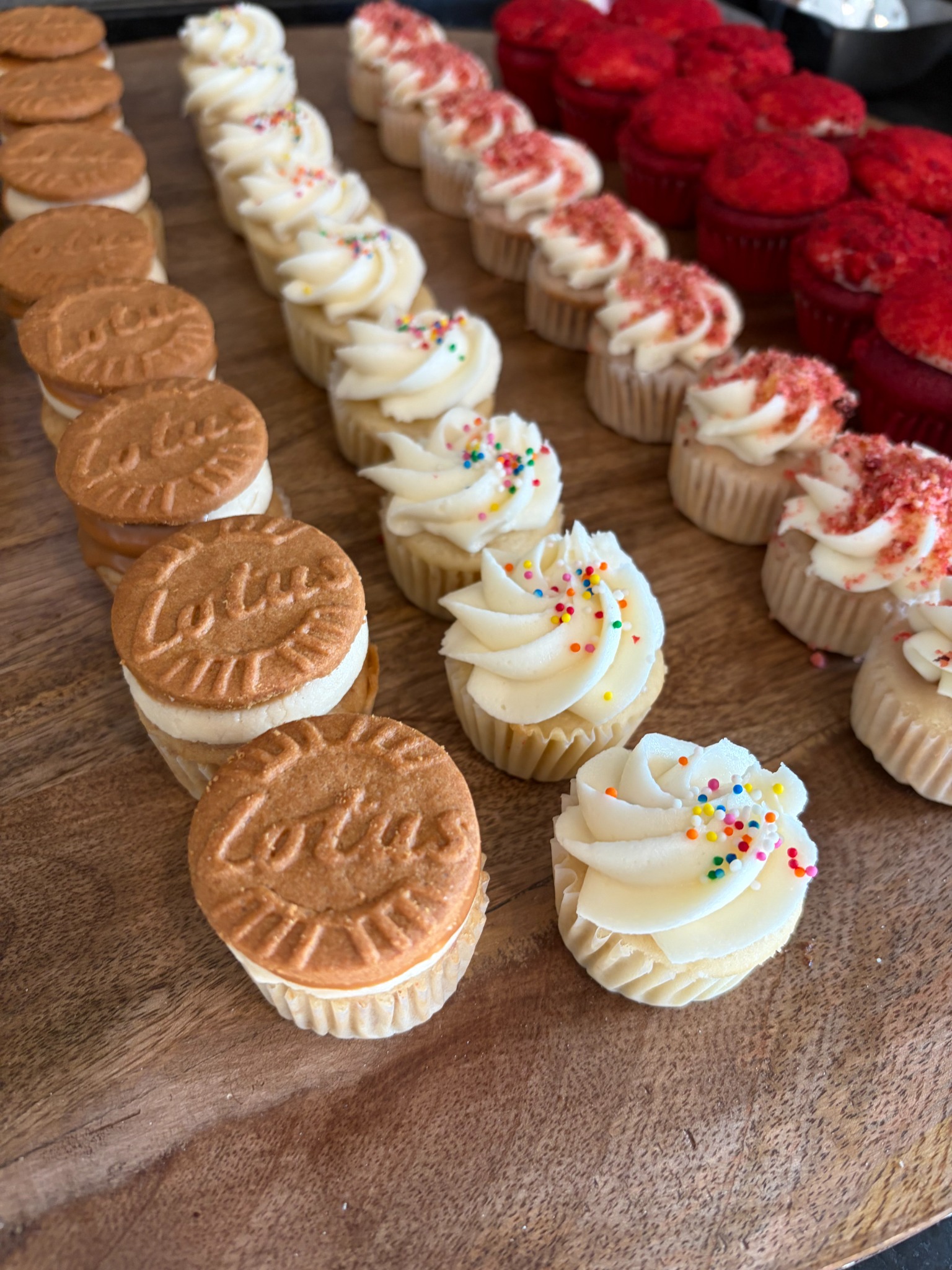 Rows of mini cupcakes topped with Biscoff cookies, vanilla frosting, strawberry crumble, and red velvet